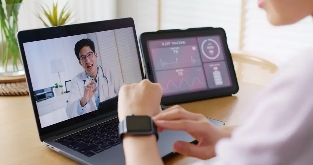 A woman wearing a smartwatch is having a telehealth consultation with a doctor on her laptop while monitoring sleep tracking data on a tablet. This image highlights the growing role of telemedicine and wearable health technology in personalized hormone health, enabling individuals to track and optimize their well-being from home.