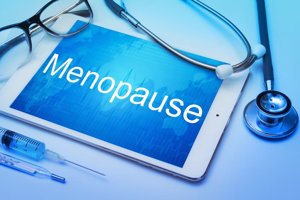 A digital tablet displaying the word "Menopause" surrounded by a stethoscope, glasses, and a syringe, symbolizing medical treatment options like Hormone Replacement Therapy.