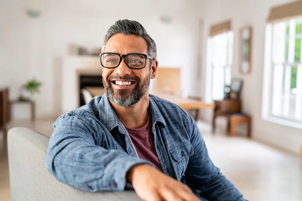 A middle-aged man with glasses and a well-groomed beard smiling confidently, representing the positive effects of hormone optimization on energy, mood, and overall well-being in a bright, modern living room.