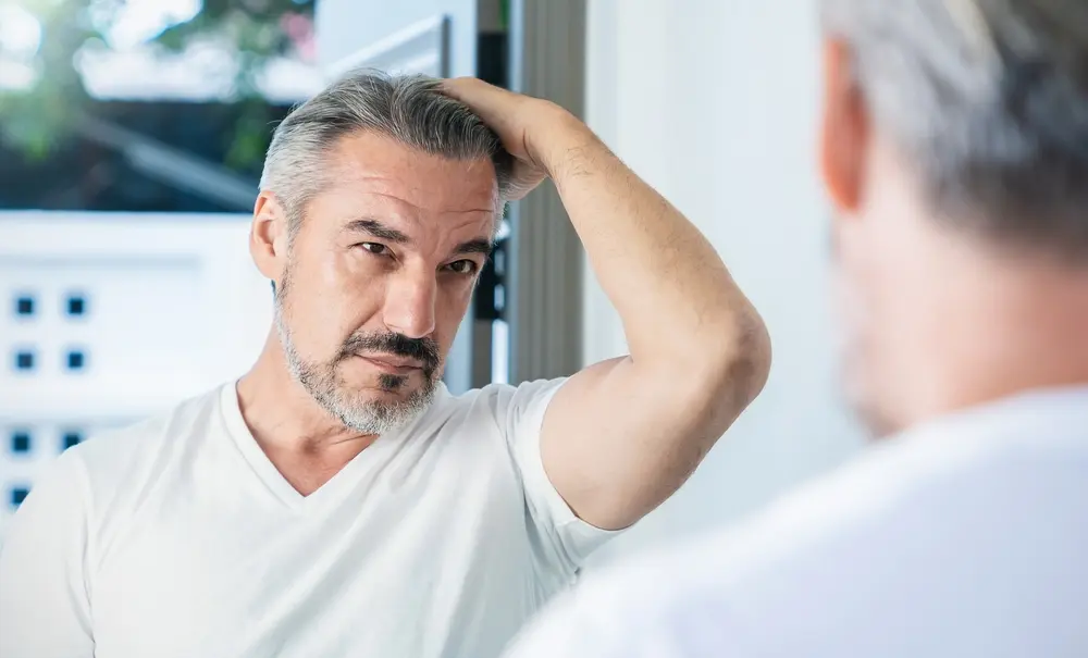Middle-aged man with graying hair examining his hairline in the mirror, concerned about hair restoration.